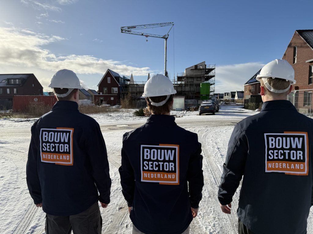 Drie vakmensen van Bouwsector Nederland met witte helm staan in de sneeuw voor een straat met huizen in aanbouw
